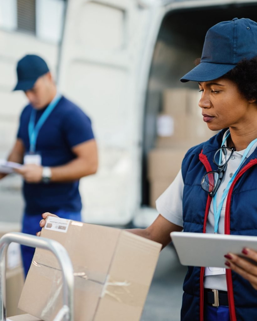 Black female worker using digital tablet while doing package inventory before the delivery. Her coworker is in the background.